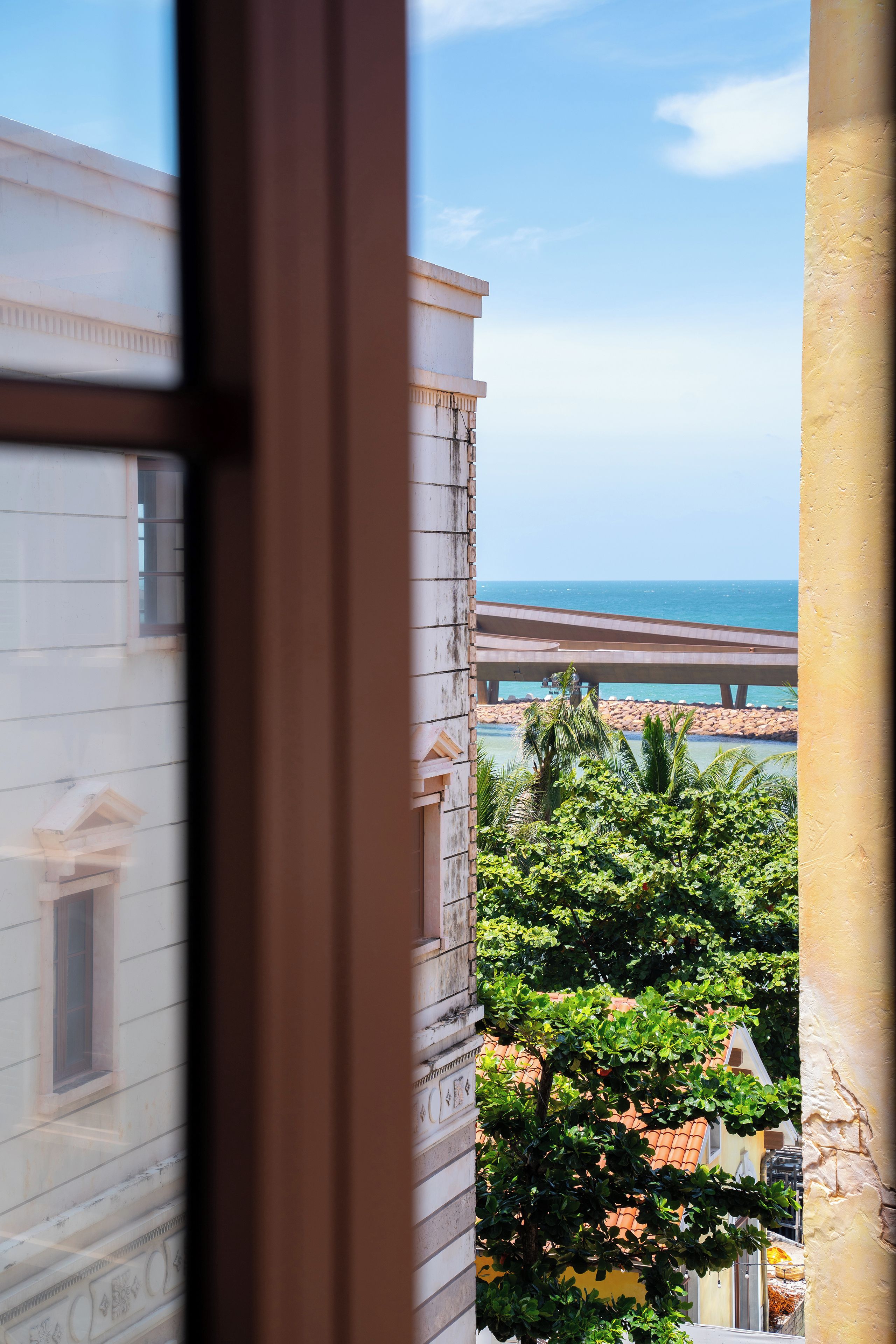 A view through a window of a building with a balcony overlooking a beach and a bridge.