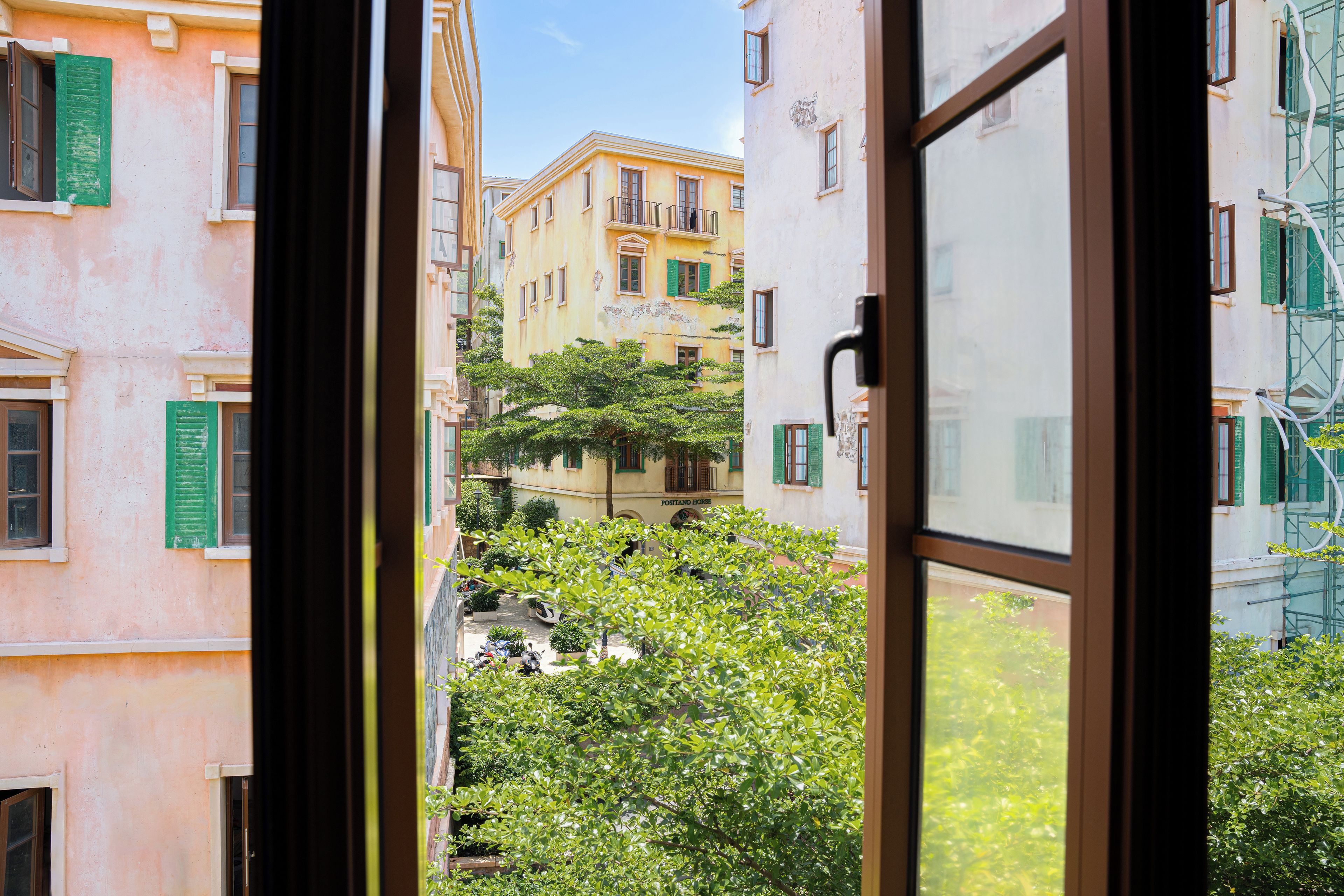 Vista a través de una ventana de un edificio de varios pisos, con balcones y un patio con árboles y scooters estacionados.