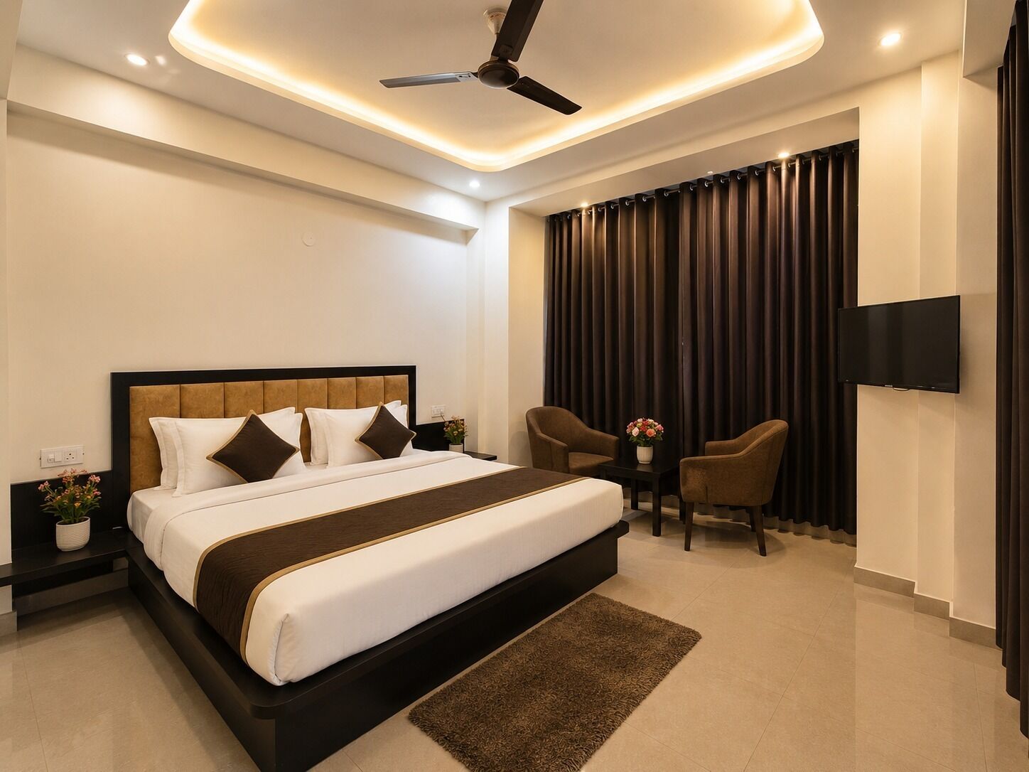 A modern hotel room with a large bed, a ceiling fan, a flat-screen TV, and a seating area with two chairs and a small table.