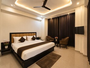 Una habitación de hotel moderna con una cama grande, ventilador de techo, televisor de pantalla plana y un área de descanso con dos sillones y una mesita.