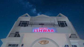 Un hotel llamado 'HOTEL ISHWAR' con un letrero llamativo, fachada simétrica y un balcón con barandilla.