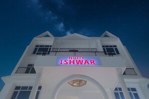 Un hotel llamado 'HOTEL ISHWAR' con un letrero llamativo, fachada simétrica y un balcón con barandilla.