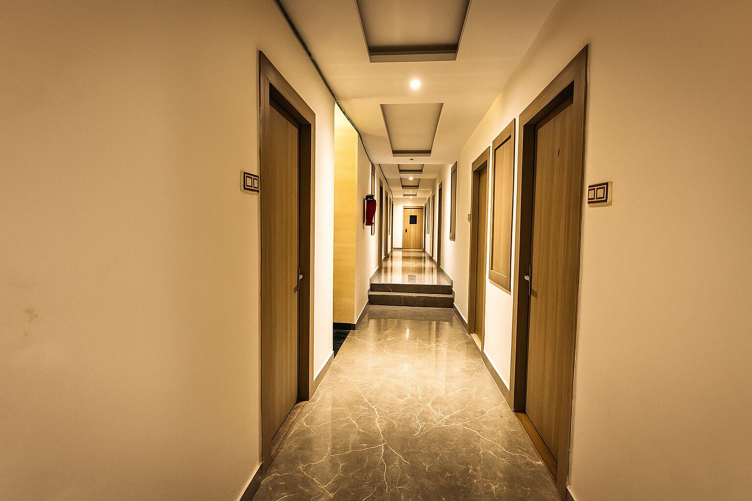 Un pasillo de hotel con puertas a ambos lados, suelo de mármol e iluminación empotrada en el techo.