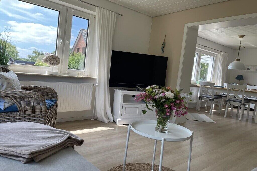 Una sala con televisor de pantalla plana, un comedor con mobiliario blanco y un ventanal con cortinas.