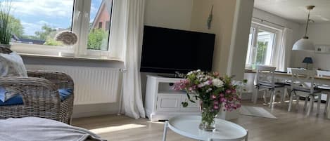 Una sala con televisor de pantalla plana, un comedor con mobiliario blanco y un ventanal con cortinas.