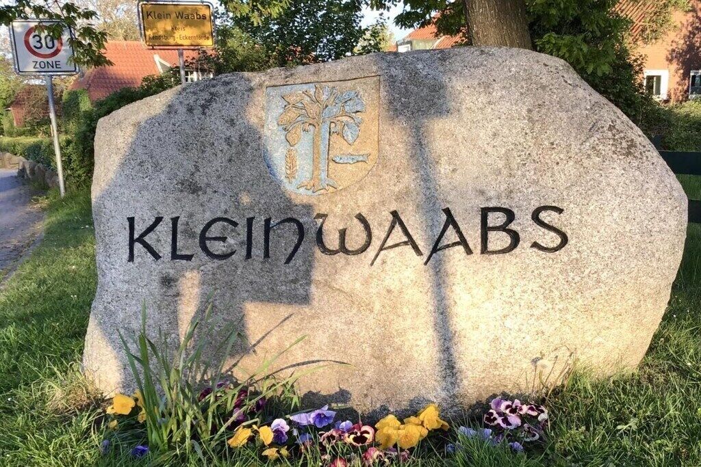 Une plaque de pierre portant le nom « Klein Waabs » et un blason, le tout entouré d’herbe et de fleurs.