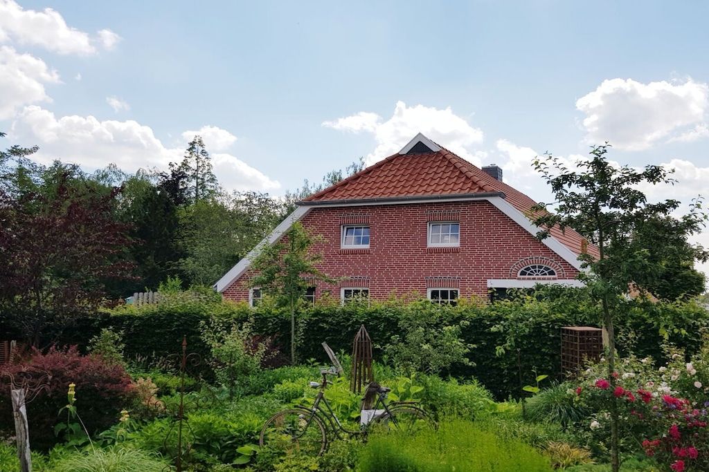 Una casa a due piani in mattoni con tetto in tegole rosse, circondata da vegetazione rigogliosa e diverse piante.