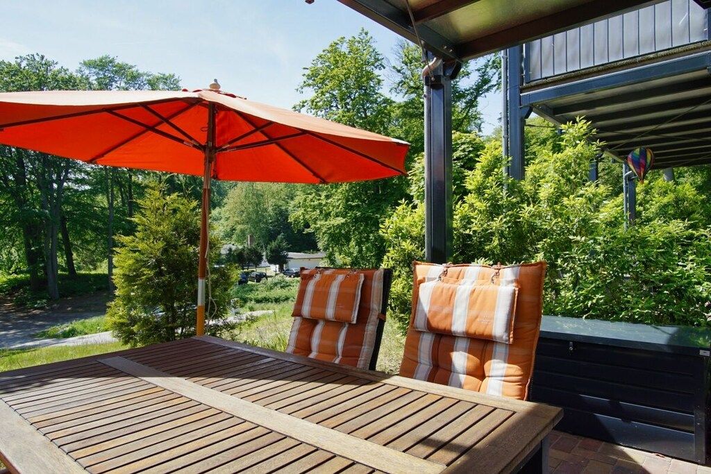 An outdoor seating area with a table and chairs, a red umbrella, and a view of trees and a building in the background.