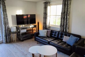 A living room with a black leather sofa, two white round tables, a flat-screen TV on a stand, and a window with curtains. - Thatched house "Aquata" (Zierow)