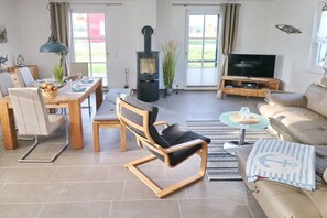 A modern living room with a sofa, a dining table with chairs, a TV, and a wood-burning stove. - Thatched house "Koje15" (Zierow)