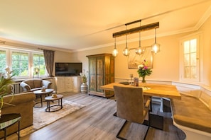 A modern living room with a dining area, wooden flooring, and a large window. - Westwind Country House (Keitum)
