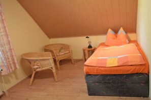 A small, cozy room with a sloped ceiling, a single bed with orange bedding, a wicker chair, and a small table with a lamp. - Holiday home with 2 bedrooms (Plau am See)