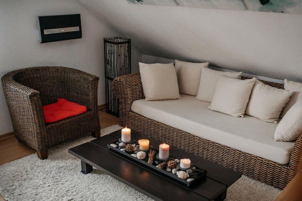 A cozy living room with a wicker sofa, a red cushion, a black coffee table with candles, and a white rug.