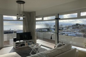 Un saló modern amb un televisor gran, un sofà i un balcó amb vista al mar i edificis.