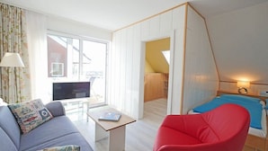 Ein kleines Zimmer mit einer Couch, einem roten Sessel, einem kleinen Tisch, einem Fernseher und einem Bett.