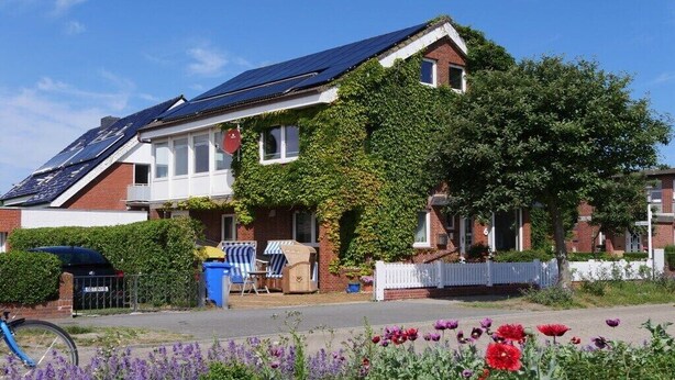 Ein Wohnhaus mit Solaranlage auf dem Dach, ein Garten mit violetten und roten Blumen und ein weißer Zaun.