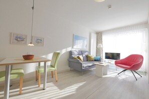Un salon moderne comprenant un coin repas, un canapé gris avec des coussins, un fauteuil rouge et un téléviseur.