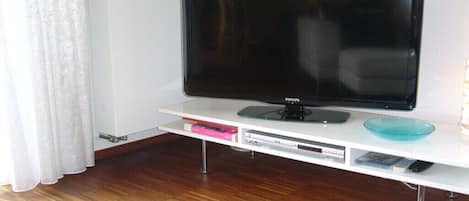 A flat-screen TV on a white entertainment unit with a remote control, a blue glass bowl, and a pink book.