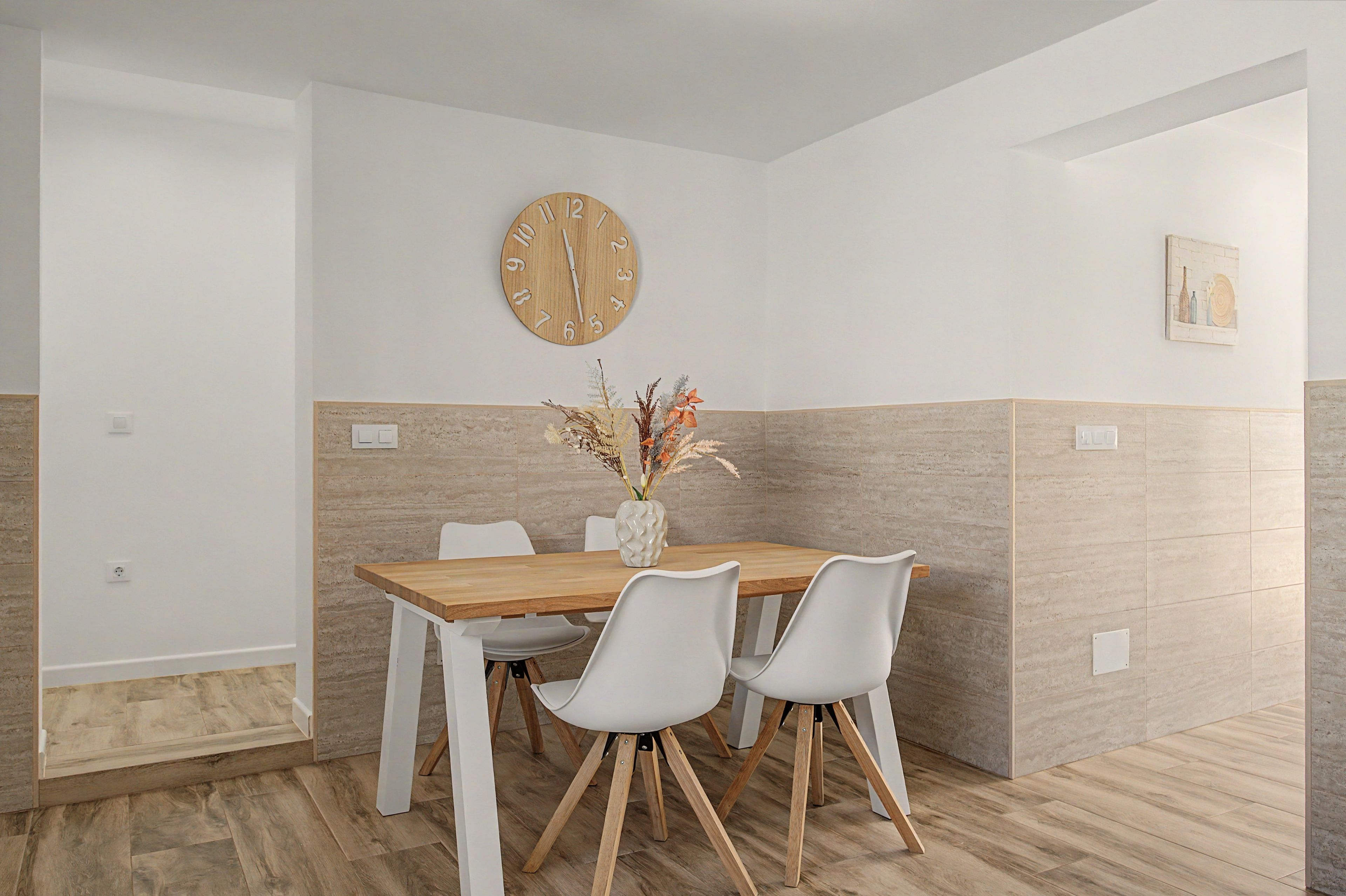 Een eethoek met een houten tafel, witte stoelen, een wandklok en een vaas met gedroogde bloemen.
