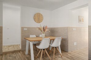 Een eethoek met een houten tafel, witte stoelen, een wandklok en een vaas met gedroogde bloemen.