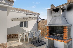Een terras buiten met een barbecue, witte stoelen en een tafel.