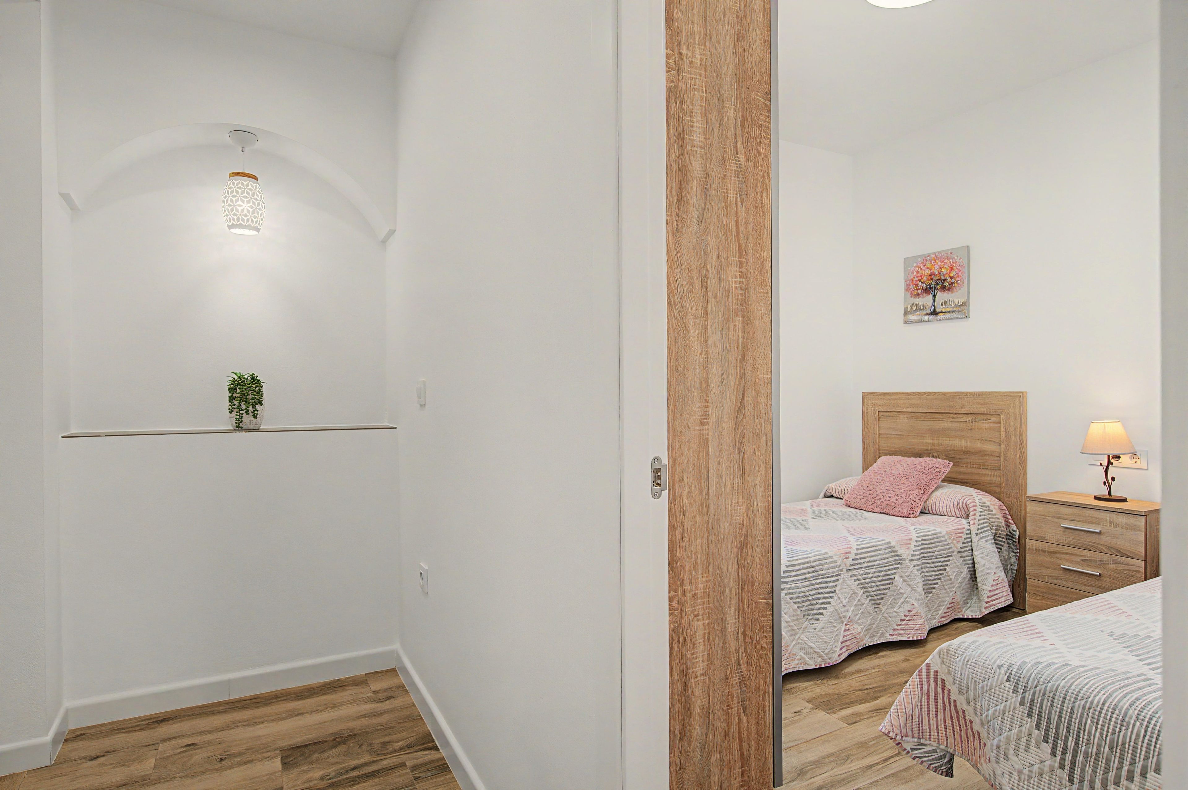Une chambre avec une tête de lit en bois, une table de chevet avec une lampe, un lit avec un oreiller rose et une étagère murale agrémentée d’une plante.