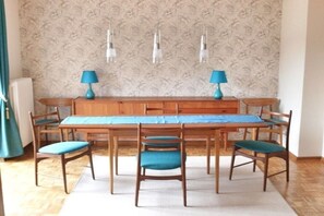 A dining area with a wooden table and chairs, a patterned wallpaper background, and pendant lights. - Dike warden - Directly in Wyk (Wyk\/Föhr)