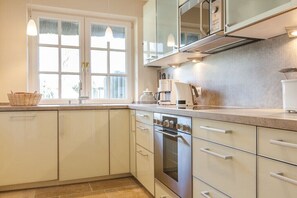 A kitchen with white cabinets, a built-in oven, and a window with curtains. - Holiday home with 3 bedrooms (Wenningstedt-Braderup)