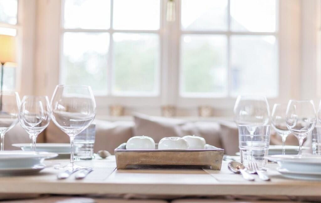 A dining table set with multiple wine glasses and plates, a tray with white spherical objects, and a window with a view of greenery.