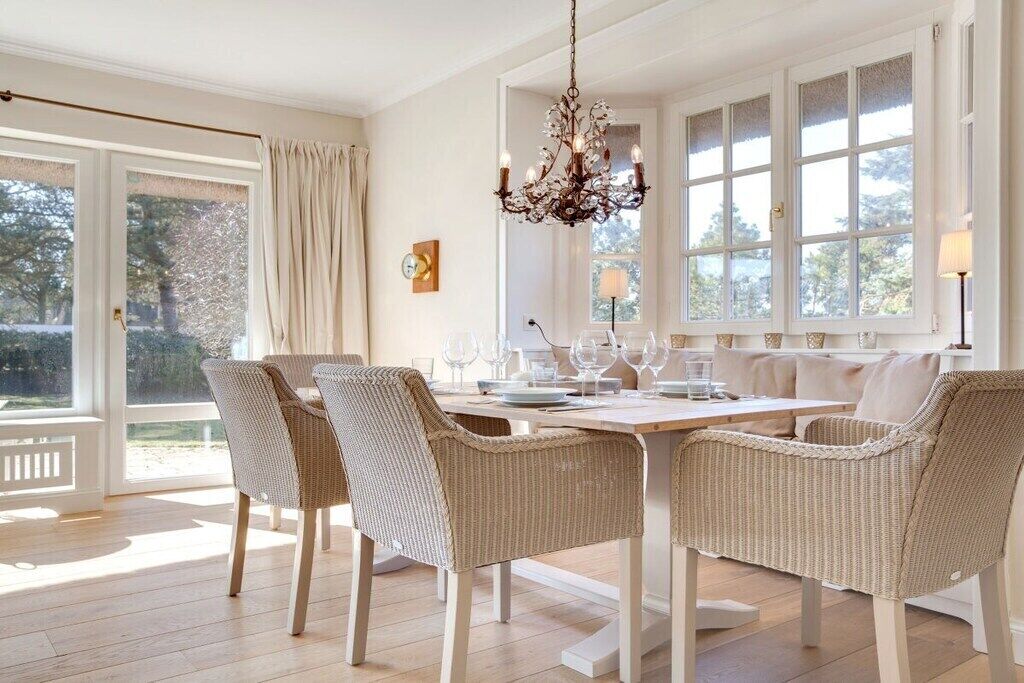 A dining area with a table set for four, surrounded by wicker chairs, a chandelier, and large windows with curtains.