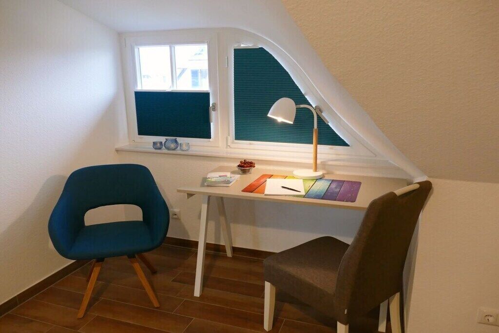 Ein kleiner, gemütlicher Raum mit schrägen Wänden, einem Schreibtisch mit Lampe, zwei Stühlen und einem Fenster mit Jalousien.