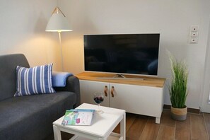 Ein Wohnzimmer mit einer Couch, einem Couchtisch mit einem Buch, einem Fernseher auf einem Schrank, einer Lampe und einer Pflanze.