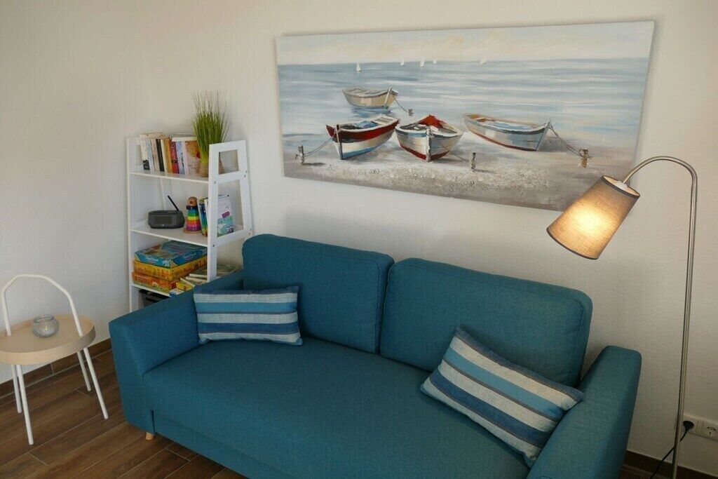 A blue sofa with striped pillows, a bookshelf with books and a plant, and a painting of boats on a beach.