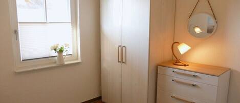 A room with a wardrobe, a bedside table with a lamp, and a window with a plant.