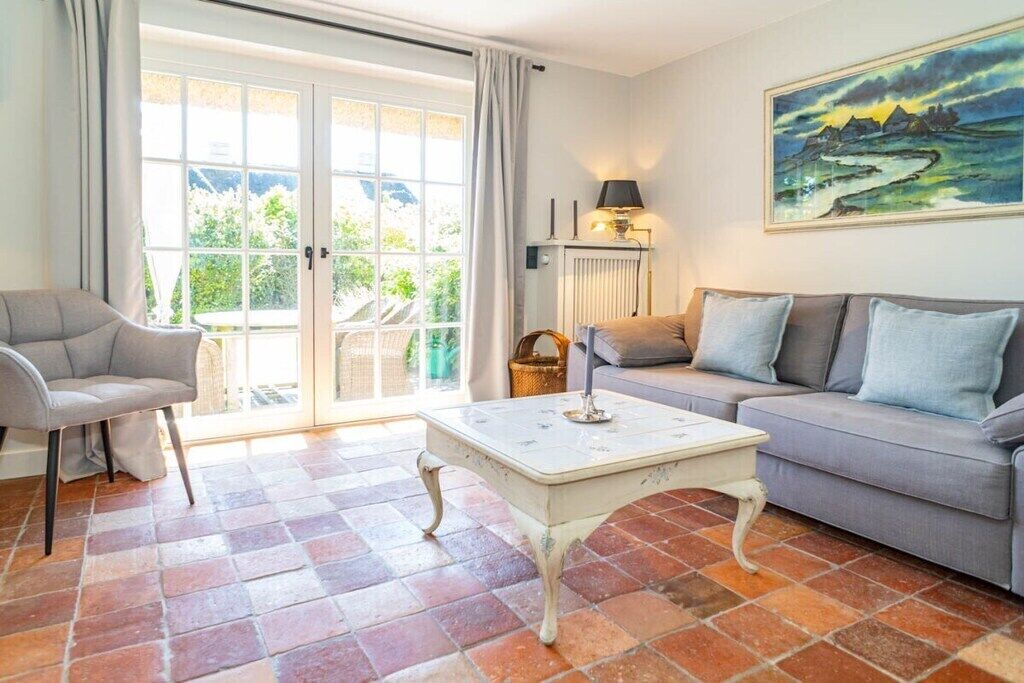 A living room with a sofa, armchair, and a table. There is a window with curtains, a radiator, and a painting on the wall.