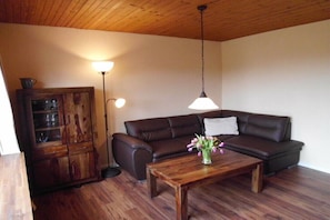 A living room with a dark brown leather sofa, a wooden coffee table with a vase of flowers, a wooden cabinet with glass doors, and a floor lamp. - Holiday home Rosenbrock (Undeloh)