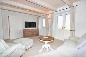 Ein Wohnzimmer mit einer Couch, einem Hocker und einem runden Tisch. An der Wand hängt ein Fernseher und es gibt ein Fenster mit Vorhängen.