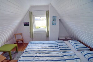 Un petit logement composé d’une seule pièce, avec un plafond en pente, un lit avec une literie à rayures, une petite table, une chaise et une fenêtre avec des rideaux.