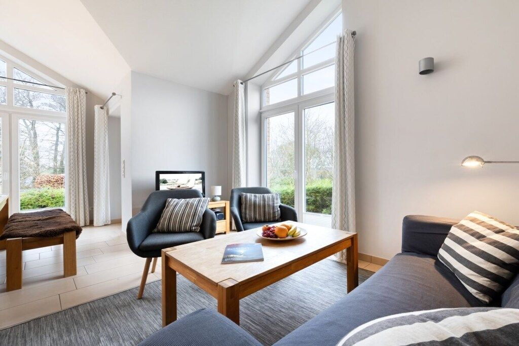 A modern living room with a sofa, a dining table, and a chair. There are large windows providing natural light.