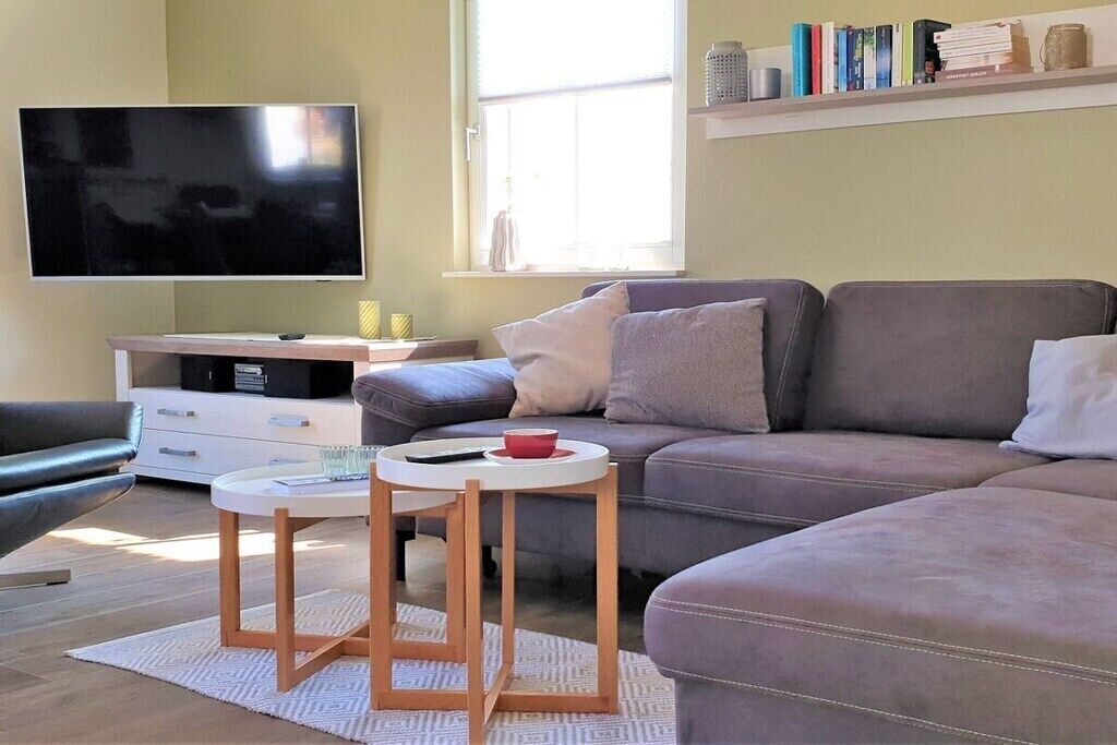 Een moderne woonkamer met een grijze bank, een flatscreen televisie, een klein rond tafeltje en een boekenkast met boeken.