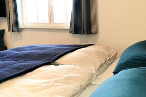 A bed with a blue blanket and pillows, a window with curtains, and a plain wall. - Thatched house "Baltic Sea shell" (Zierow)