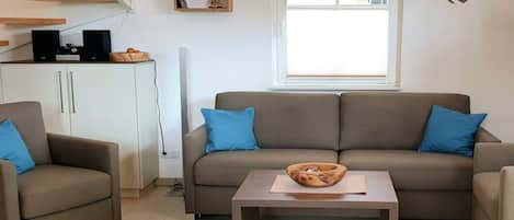 Een woonkamer met een bank, twee fauteuils, een salontafel, een witte kast en een raam met jaloezieën.