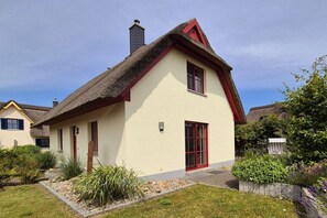 A traditional house with a thatched roof, a red door, and a small garden with various plants. - Thatched house "Baltic Sea shell" (Zierow)
