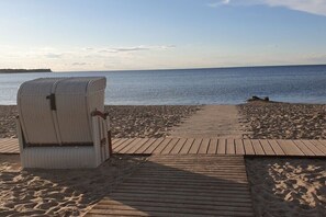 A beach with a wooden boardwalk leading to a white wicker chair. - Holiday home in Zierow with swimming pool (Zierow)