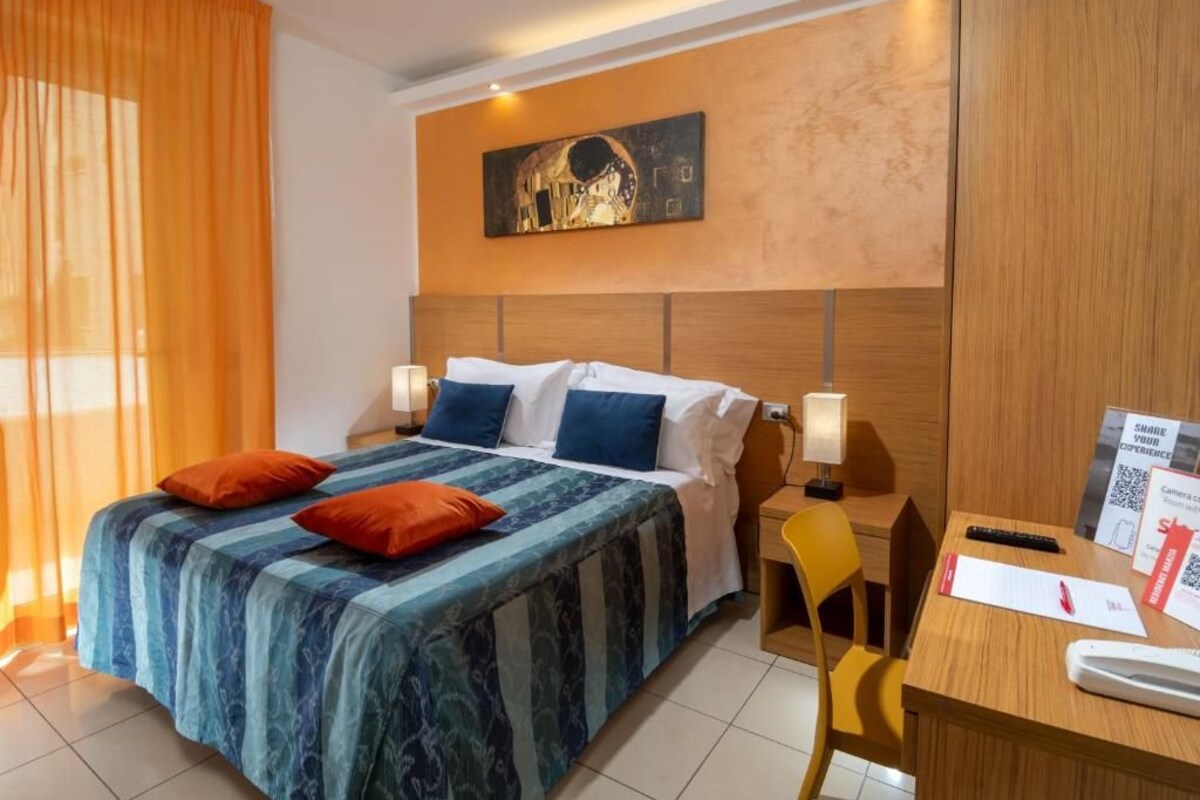 A hotel room with a bed, two pillows, a desk with a chair, a wall-mounted artwork, and a sign with a QR code.