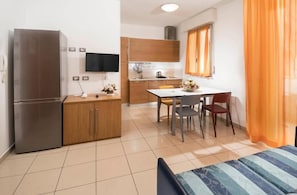 A compact kitchen with a refrigerator, wooden cabinets, a dining table with chairs, and a TV mounted on the wall. - Residence Marzia (Rivazzurra)
