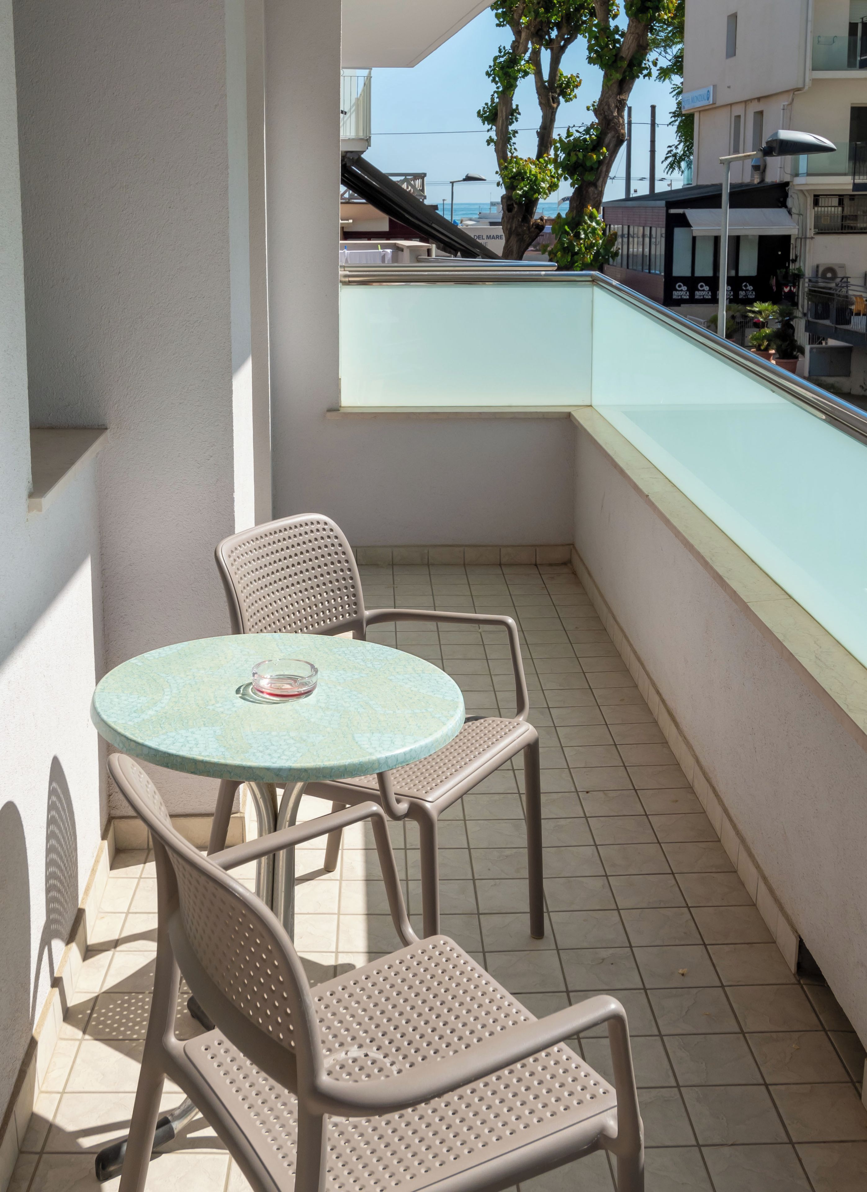 Un balcón con una mesa redonda y dos sillas, piso de baldosas y vista al mar y edificios al fondo.