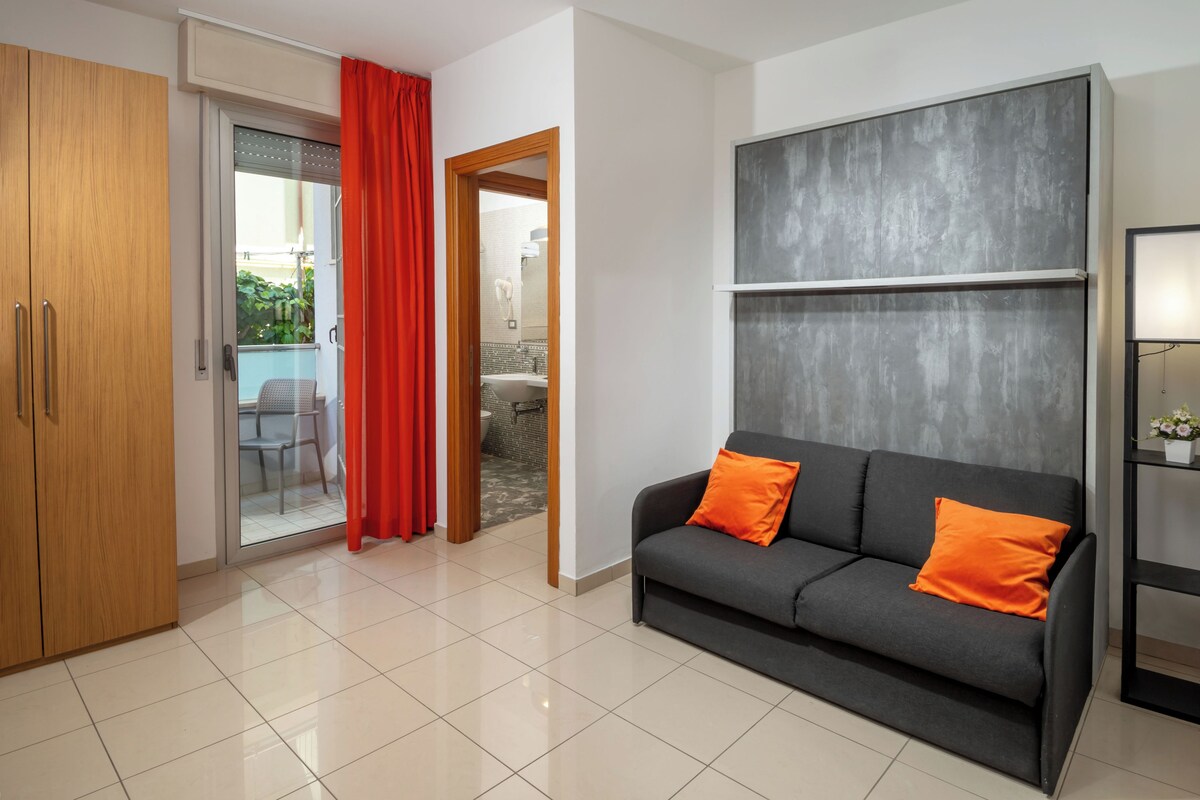 A modern living room with a grey sofa, red cushions, a wooden wardrobe, and a sliding glass door leading to a balcony.