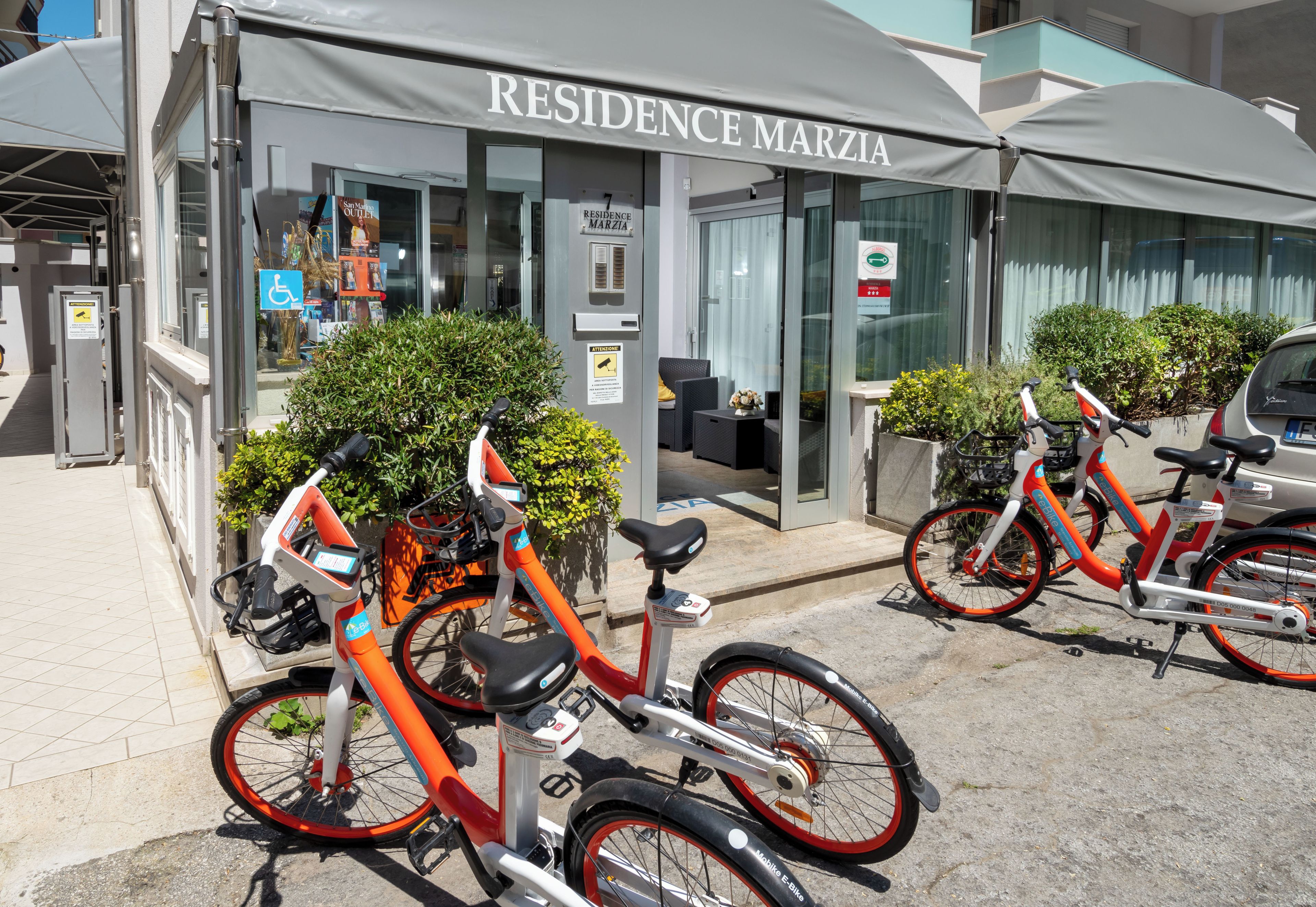 A residential building named 'Residence Marzia' with a covered entrance, a bicycle rental station, and a parking area.