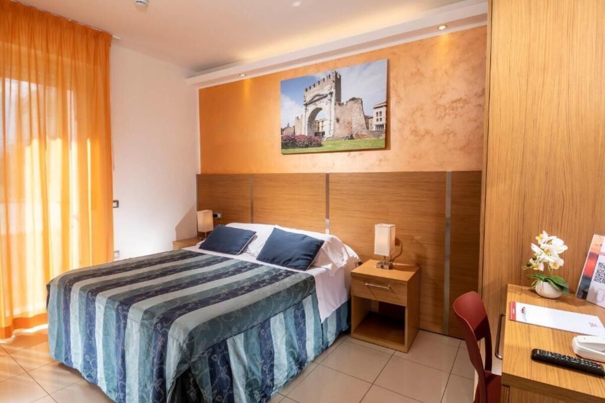 A hotel room with a bed, a desk, a chair, a nightstand, and a painting on the wall.